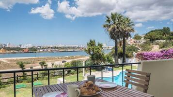 A balcony with a table set for breakfast, overlooking a coastal cityscape and a swimming pool.