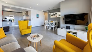 Una sala de estar moderna con un sofá, sillones amarillos, una mesa de centro y un comedor con sillas y un televisor.