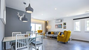 Una sala de estar moderna con un sofá gris, sillones amarillos y una mesa de comedor de vidrio con sillas blancas.
