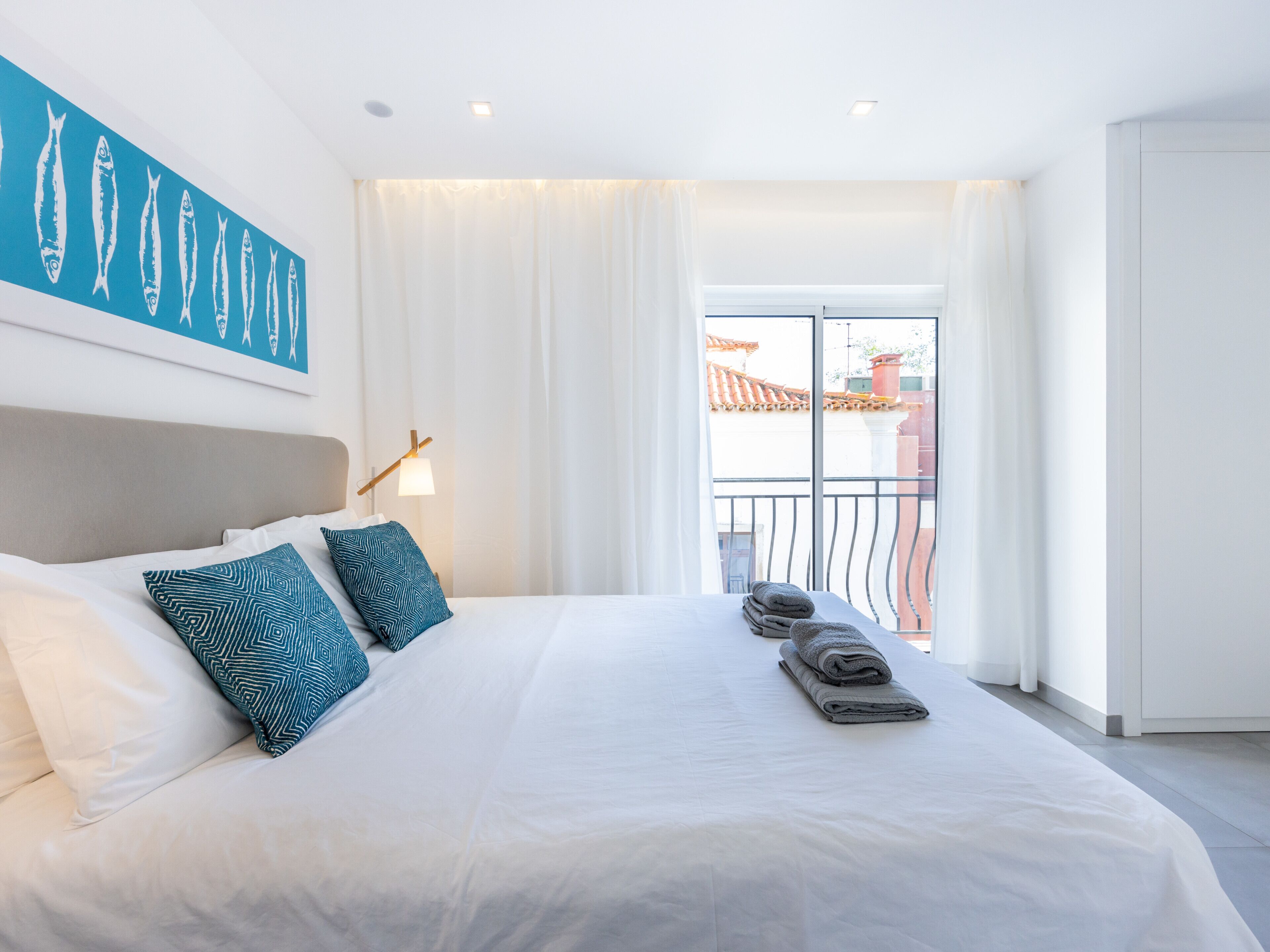 Ein Hotelzimmer mit einem großen Bett, weißen Bettwäsche, zwei gemusterten Kissen, einer Wandlampe und Blick auf einen Balkon mit Geländer.