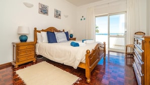 A bedroom with a wooden bed, a nightstand, a dresser, and a view of the sea through a large window. - Sunny Seaside Escape (Lagos)