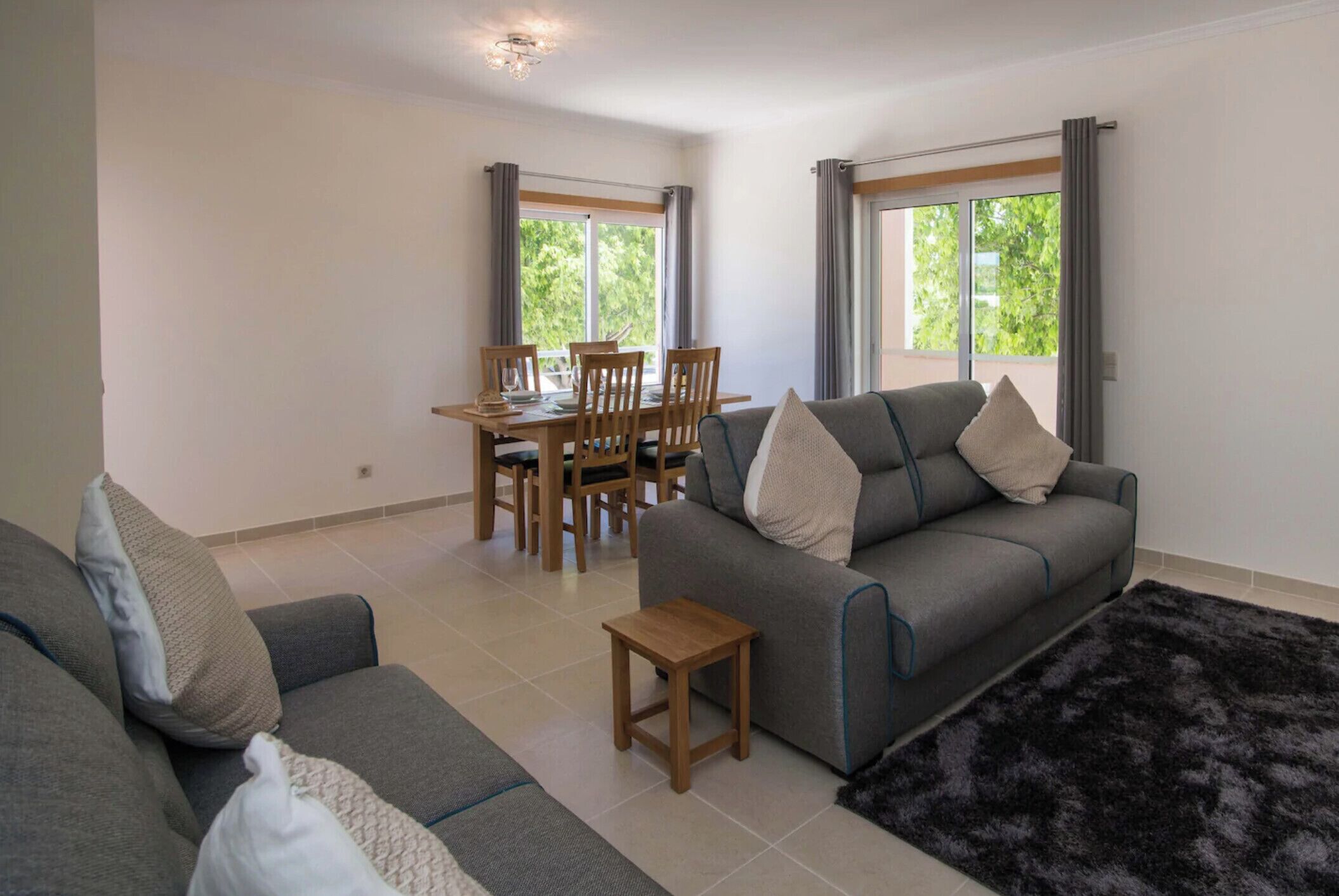 A modern living room with a grey sofa, a small wooden table, and a black furry rug. There is a dining area with a wooden table and chairs, and large windows with curtains.
