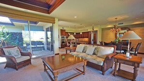 A spacious living room with a large sofa, two armchairs, and a coffee table. A kitchen with dark wooden cabinets and a dining area with a table and chairs are visible in the background. - Home Away From Home - Beautiful Townhome in the Heart of Mauna Lani (Kamuela)