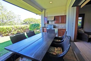 Un'area pranzo all'aperto con un lungo tavolo e sedie, una cucina sullo sfondo e vista su un giardino.