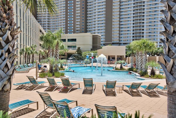 A resort pool area with lounge chairs, a central water feature, and tall buildings in the background.