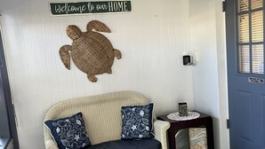 A wicker sofa with dark cushions, a welcome sign, a decorative turtle, a small table with a glass top, and a blue door. -  Downtown Safety Harbor 2 Bed 1 Bath Pool Home, 20 minutes from Clearwater beach (Safety Harbor)