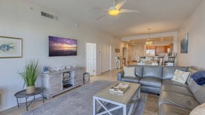 A spacious living room with a grey leather sofa, a coffee table, and a TV mounted on the wall. There is a dining area visible in the background with wooden cabinets and a chandelier.