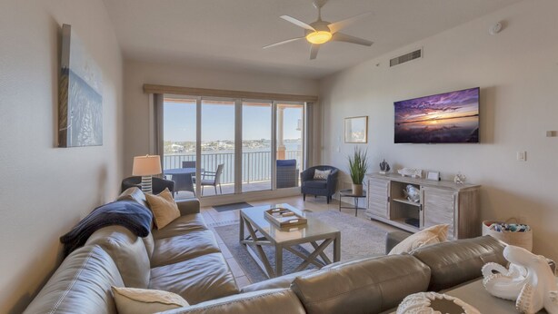 A spacious living room with a large sofa, a coffee table, and a TV mounted on the wall. There is a balcony with a view of the city and a ceiling fan.