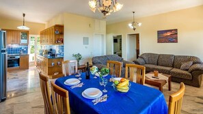 A dining area with a blue tablecloth, wooden chairs, and a fruit bowl. There is a kitchen in the background with wooden cabinets and a refrigerator.