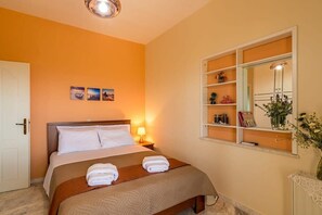 A hotel room with a bed, a bedside table with a lamp, a window with shelves, and a door.