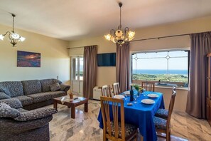 A living room with a sofa, dining table, and chairs, a chandelier, and a view of the sea.
