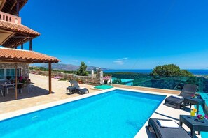 A swimming pool with lounge chairs and a table, surrounded by a stone patio and a view of the sea.