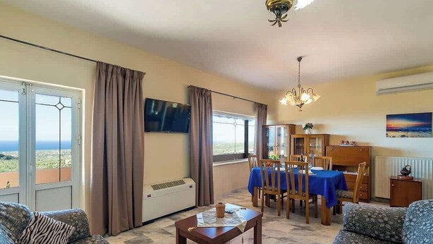 Una sala de estar con un sofá, una mesa de centro, un comedor con mesa y sillas, un televisor montado en la pared y vistas al exterior a través de amplios ventanales.
