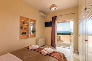 A bedroom with a bed, a shelf with decorative items, an air conditioning unit, a television, and a view of the outdoors. - Panorama Sea Breeze - Pool Dream Gem (Polemarchi)