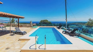 Una piscina rectangular con dos escaleras metálicas, rodeada de sillas de descanso y un patio de piedra.