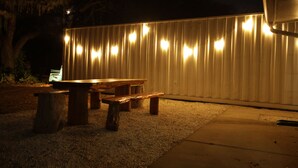 Een terras buiten met een houten tafel en banken, een metalen hek en lichtslingers.