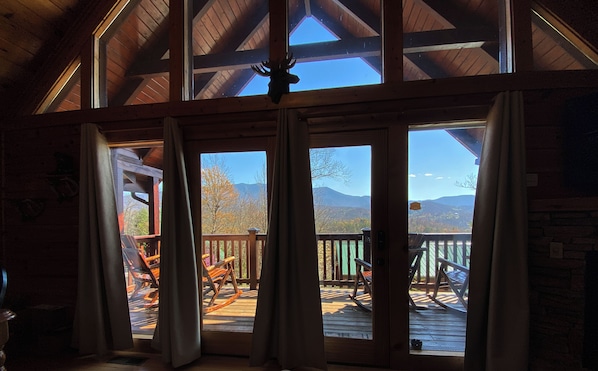 Un salón amplio con un gran ventanal corredera que da a una terraza. La habitación cuenta con vigas de madera en el techo y una chimenea de piedra.