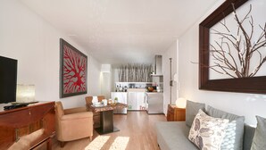 Un salon moderne avec un canapé, une table basse et un téléviseur. Au fond, on aperçoit une cuisine équipée d’un réfrigérateur et d’un coin repas.