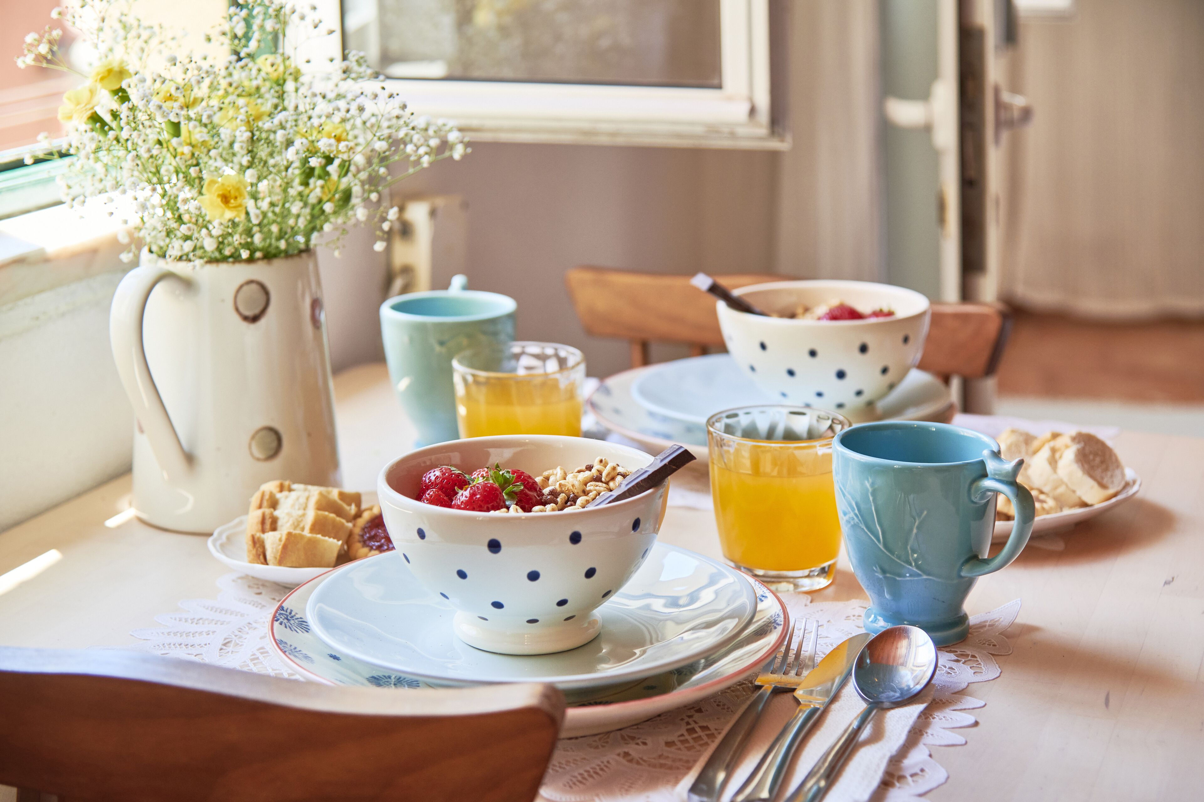 Une table de petit-déjeuner avec un vase de fleurs, un bol de céréales aux fraises, un verre de jus d’orange et une tasse bleue.