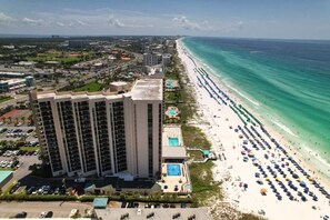 A high-rise hotel with a pool and beach view, adjacent to a long stretch of beach with umbrellas. - The Scallop | Beachfront Condo (Destin)