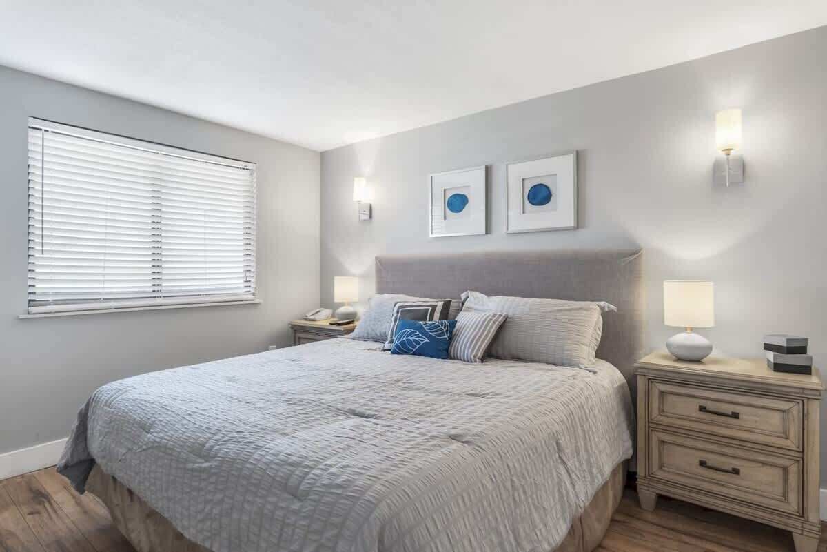 A bedroom with a bed, bedside tables, a nightstand, a window with blinds, and wall-mounted lights.