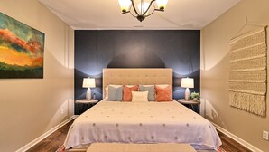 A bedroom with a large bed, two bedside tables with lamps, a wall-mounted tapestry, and a painting on the wall.