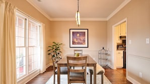 A dining area with a wooden table and chairs, a hanging light fixture, a framed picture on the wall, and a kitchen in the background.