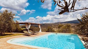 A rectangular swimming pool with a tiled deck, a pergola with a roof, and a view of a mountainous landscape.