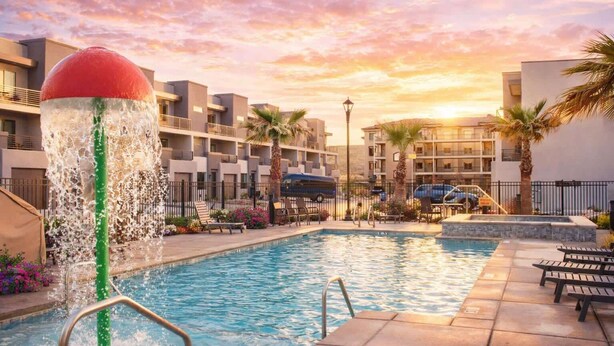 A swimming pool with a water feature, surrounded by palm trees and lounge chairs, with a backdrop of modern buildings and a sunset sky. - V 31| 3-Level Townhome *Pool* Hot Tub * Pickleball (St. George)