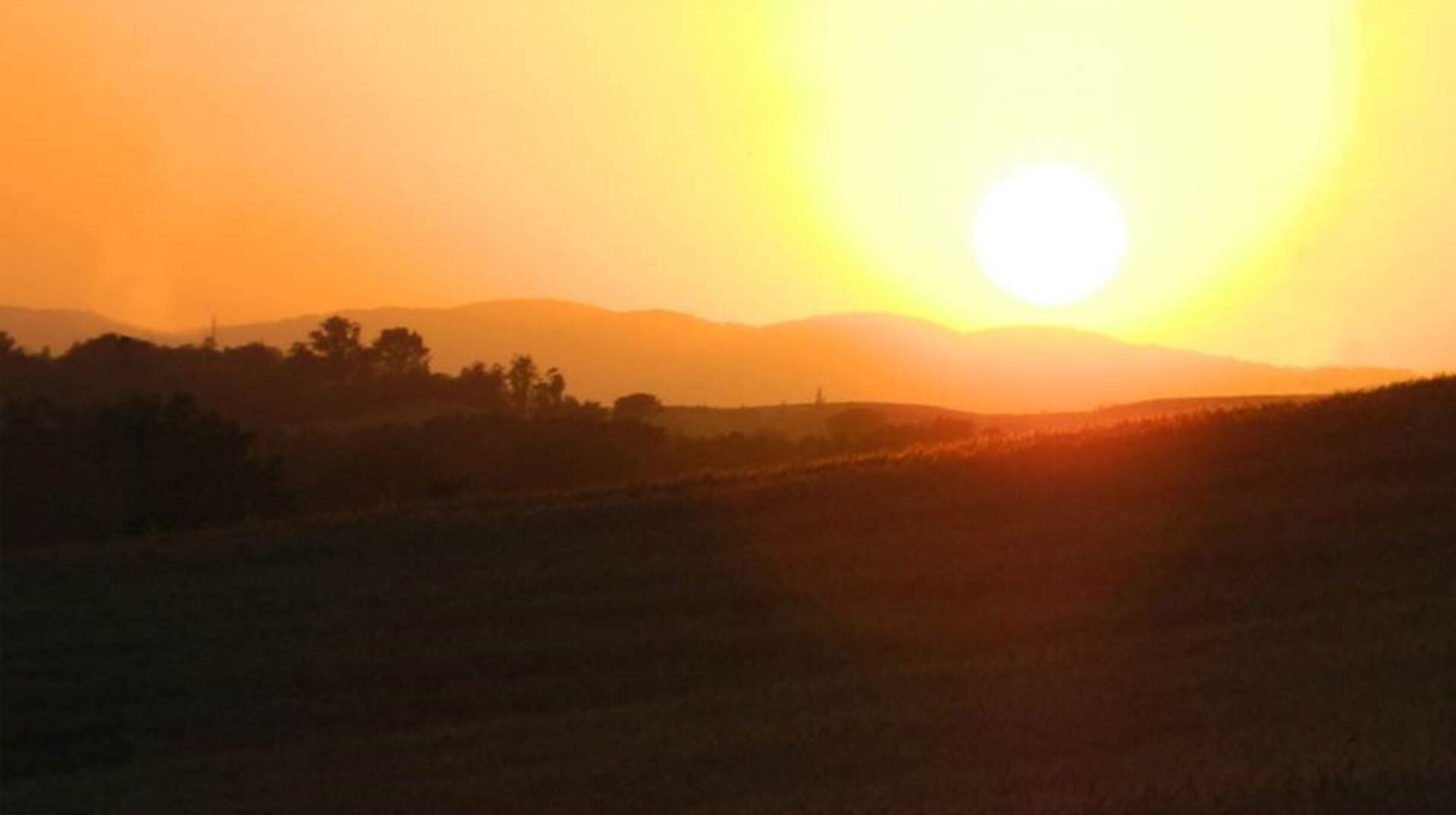 A sunset over a hilly landscape with silhouettes of trees and a distant structure.