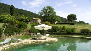A pool area with lounge chairs and an umbrella, surrounded by lush greenery and a stone building in the background. - Case Belle, beautiful villa in the heart of the Val d'Orcia with 12 sleeping accommodations (Sarteano)