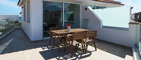 A modern outdoor patio with a table and chairs, a sliding glass door, and a view of the sea.