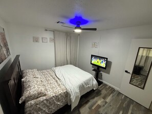 Quarto com cama, televisão, ventoinha no teto e porta.