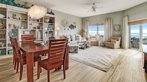 A spacious living room with a dining area, large windows with curtains, a chandelier, and a view of the ocean. - Gorgeous Ocean Views | Ocean Park Penthouse | Garage Access | Easy Walk to Beach (Fernandina Beach)