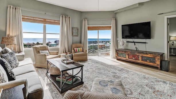 A spacious living room with a large flat-screen TV, comfortable seating, a coffee table, and a view of the ocean through large windows. - Gorgeous Ocean Views | Ocean Park Penthouse | Garage Access | Easy Walk to Beach (Fernandina Beach)