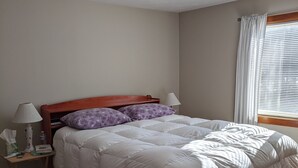 Ein ordentlich bezogenes Bett mit einem Holz-Kopfteil, ein Nachttisch mit Lampe und ein Fenster mit Vorhängen.