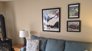 Ein Wohnzimmer mit einem blauen Sofa, einem Holztisch mit Fernbedienungen und Zeitschriften, einem schwarzen Kamin und zwei gerahmten Postern an der Wand.