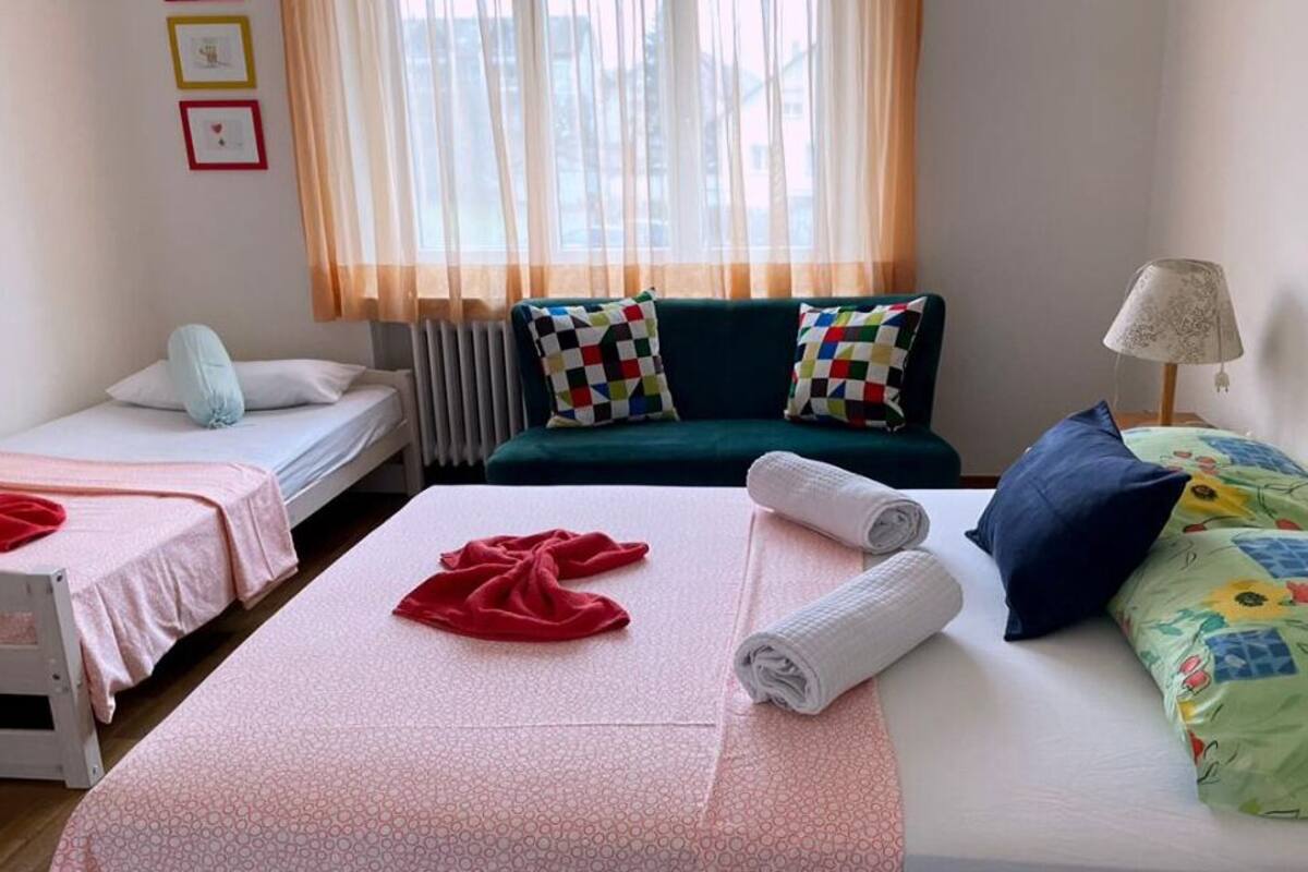 A hotel room with two beds, a sofa, a lamp, and a window with sheer curtains.