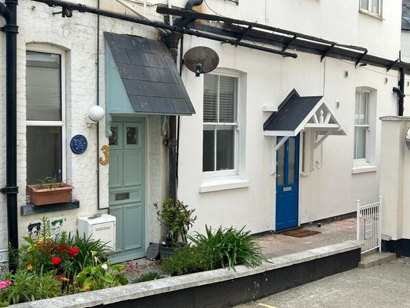 Una casa de estilo urbano blanca con una puerta azul, una puerta verde y un pequeño jardín con macetas y flores.