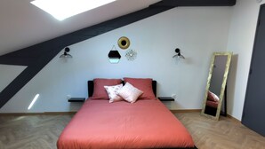 Une chambre avec un plafond en pente, un lit recouvert d’une couverture rouge, deux lampes fixées au mur, un miroir rond et une petite étagère.
