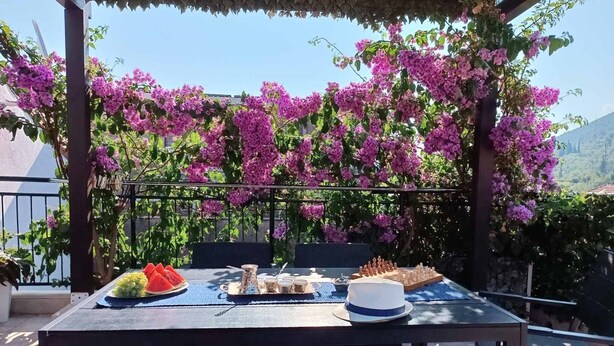 Comedor al aire libre con una mesa preparada para una comida, rodeado de flores moradas y vistas del paisaje.