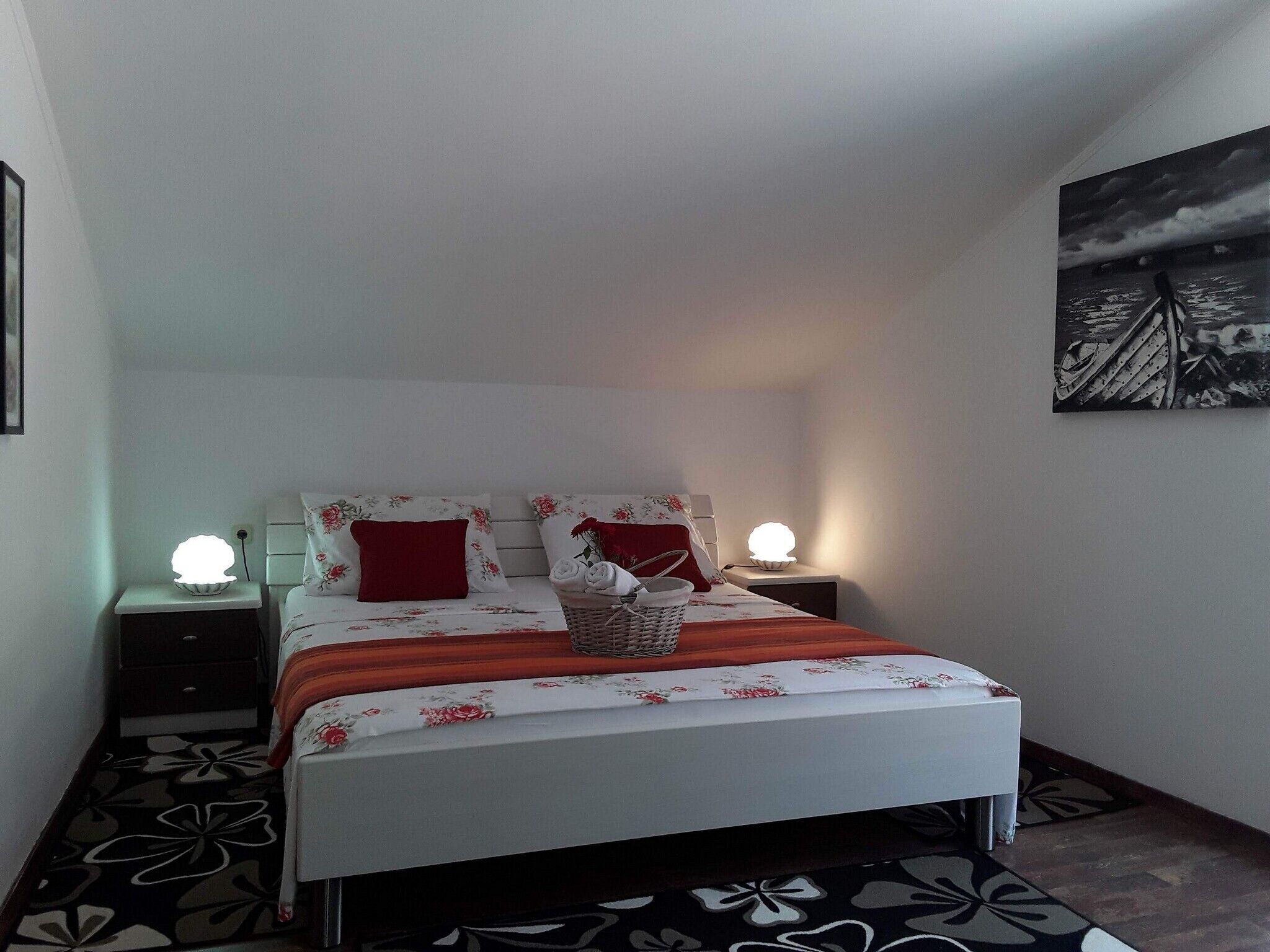 A neatly made bed with floral bedding, a red pillow, and a basket of towels. There are two bedside lamps and a wall-mounted artwork.