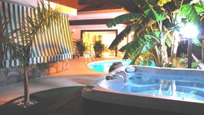 A swimming pool area with a hot tub, surrounded by tropical plants and a striped awning.