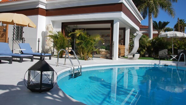 A swimming pool with lounge chairs and umbrellas, surrounded by palm trees and a building with a tiled roof.