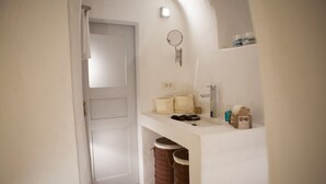 A compact bathroom with a white door, a small shelf with toiletries, a towel rack, and a trash bin. - Santorini Finikia Oia Cave House (Santorini)
