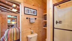 Une salle de bain rustique, aménagée dans une cabane en bois, avec une douche, des toilettes et un lit recouvert d’une courtepointe à rayures.