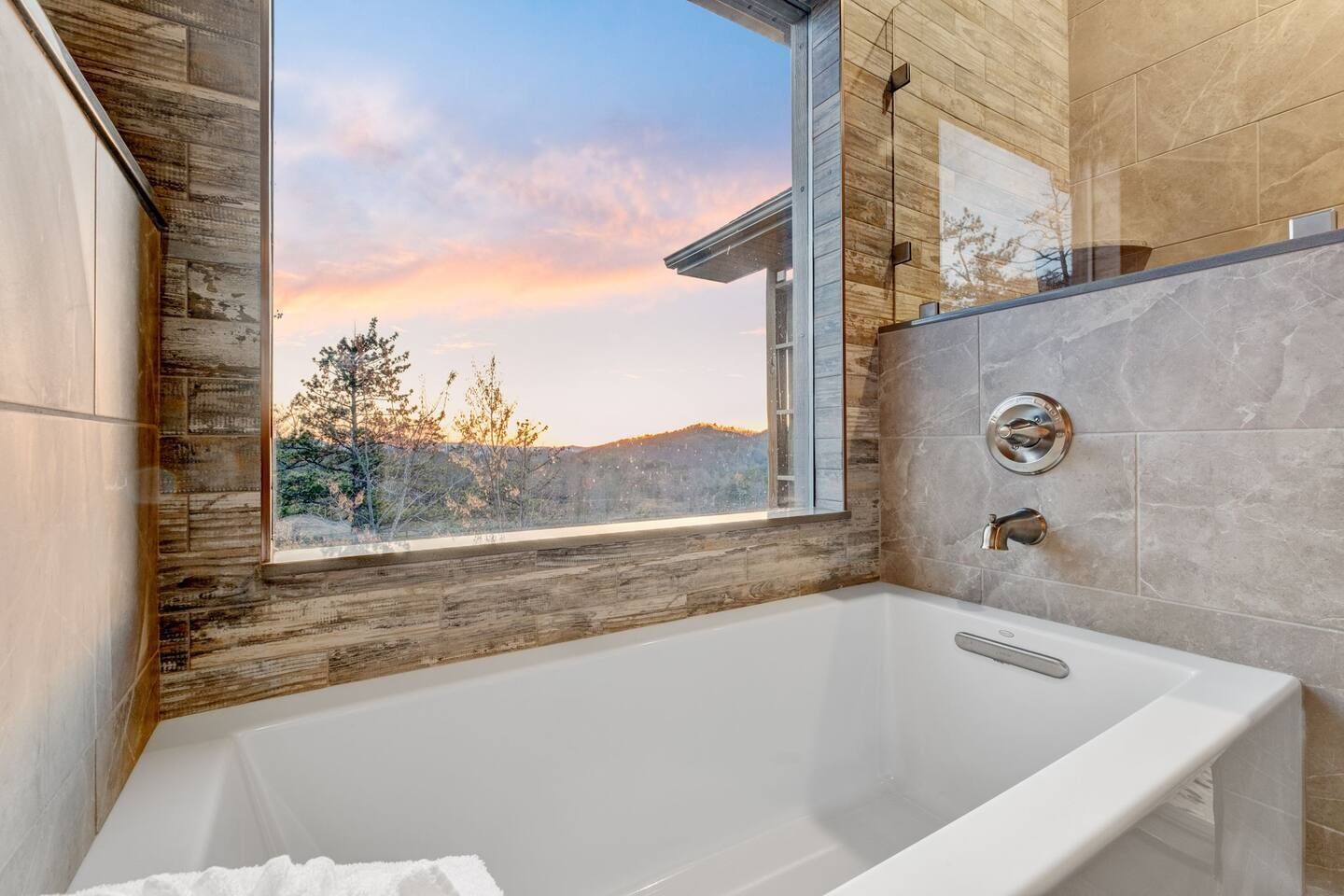 Un baño moderno con una bañera grande, una ventana que ofrece vistas a árboles y colinas, y una pared de acento de madera.