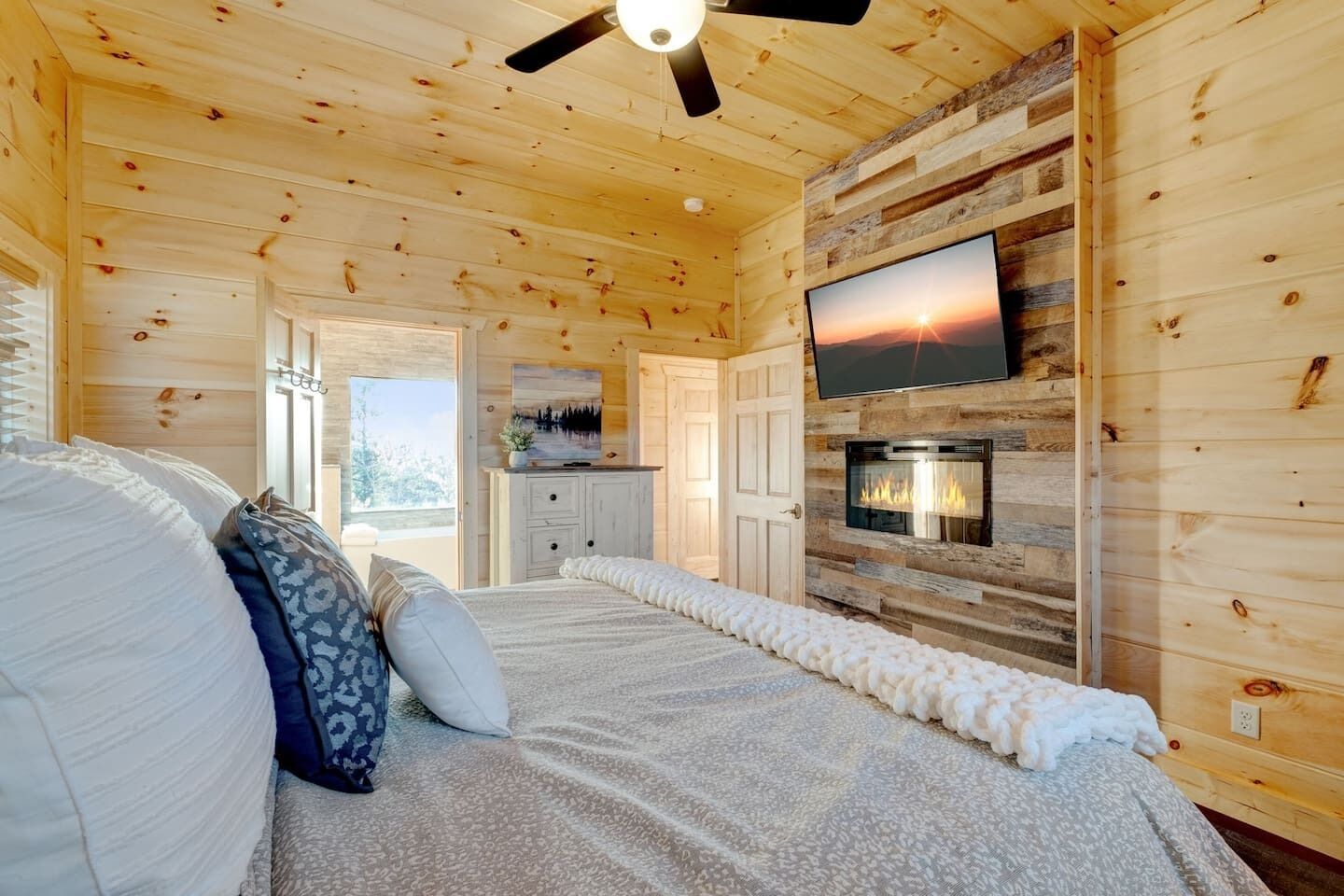 Acogedor dormitorio de cabaña de madera con cama, almohadas, chimenea, televisor y vistas al exterior.
