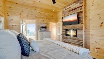 Acogedor dormitorio de cabaña de madera con cama, almohadas, chimenea, televisor y vistas al exterior.