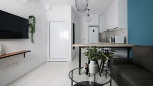 Ett modernt vardagsrum med en platt-TV, en soffa, ett soffbord med en vas och en flaska, samt en köksdel med en mikrovågsugn och en hängande lampa.